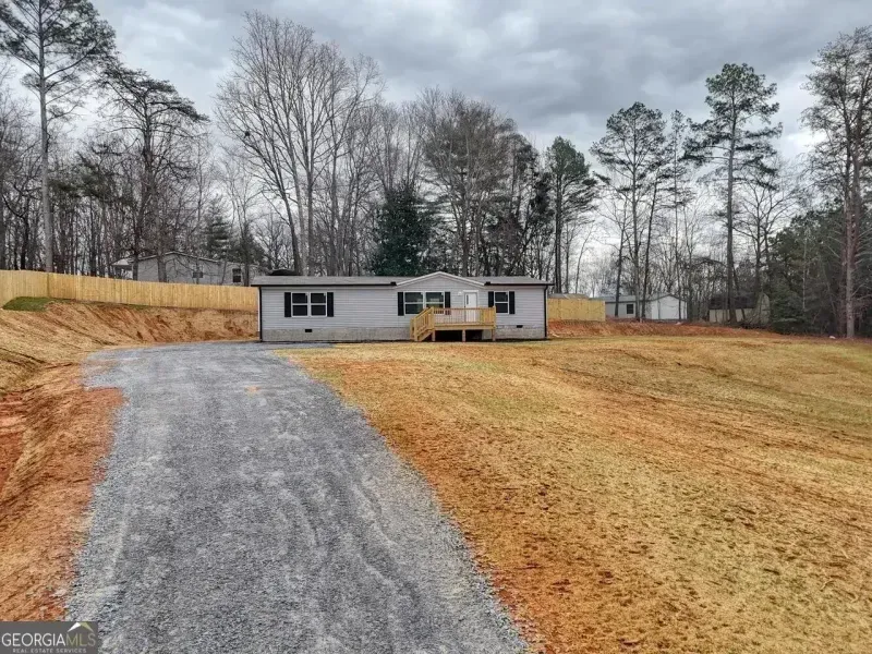 A single-story gray mobile home with a small front porch, set on a grassy lot with a gravel driveway and tree line.