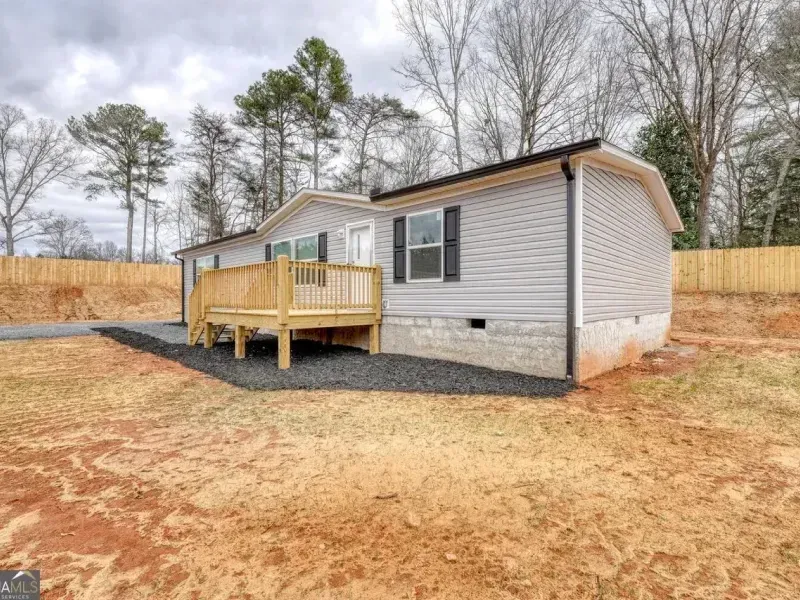 A light gray modular home with a wooden porch on a gravel pad, set on a plot of land with bare dirt and a wood fence.