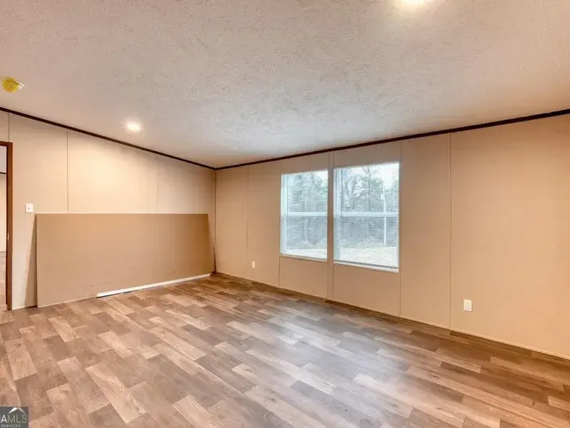 An empty room with tan walls, light wood-look flooring, two windows, and an unpainted wall panel leaning against the side.
