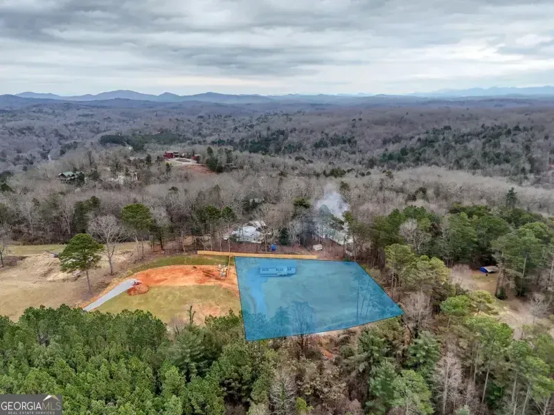 Aerial view of a cleared, blue-highlighted plot of land surrounded by forest and mountains under a cloudy sky.