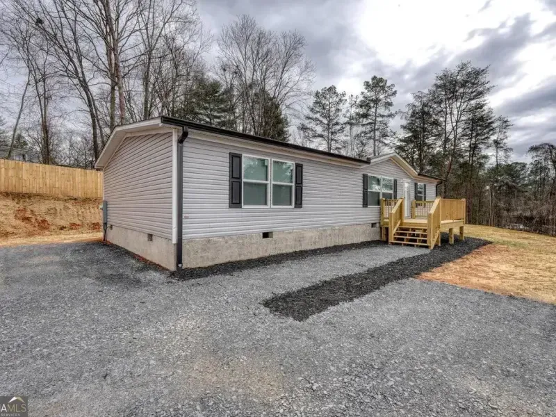 A light gray manufactured home with a tan stone foundation and a small wooden porch under a cloudy sky.