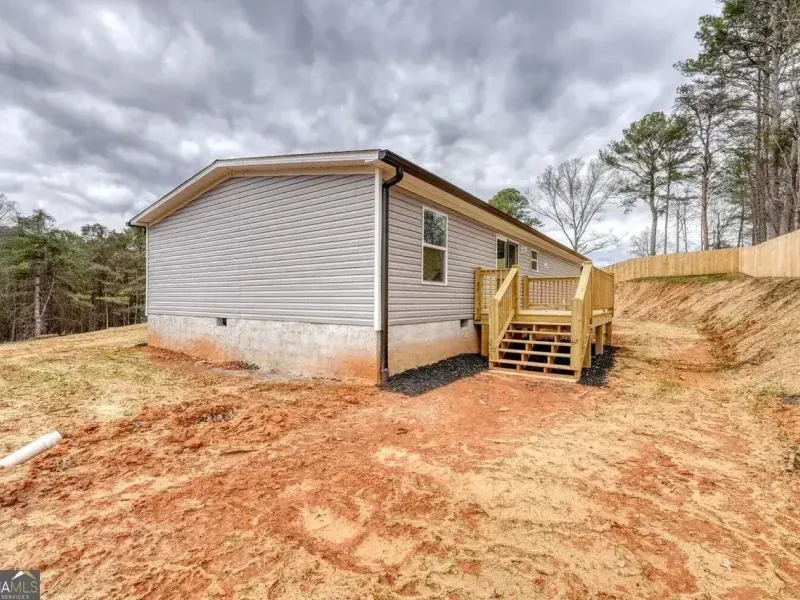 A light gray mobile home on a dirt lot with a wooden deck and staircase under a cloudy sky.