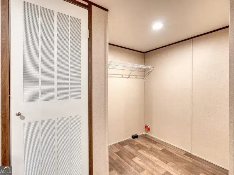 A louvered white door stands next to an empty walk-in closet with beige walls, a wire shelf, and wood-look flooring.