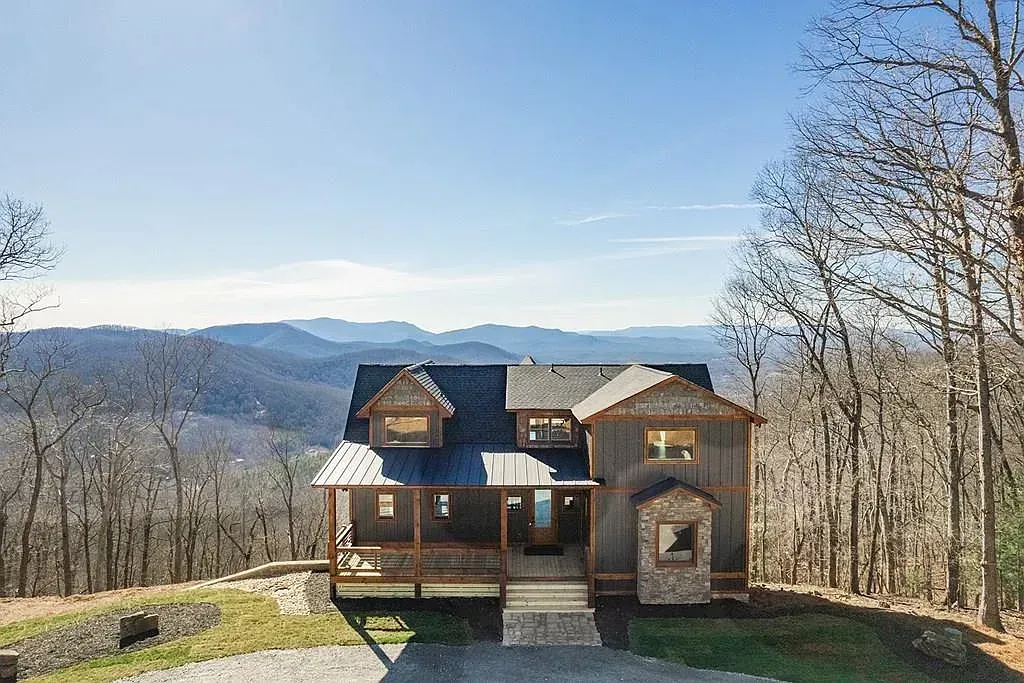 A multi-story wooden mountain home with stone accents and a metal roof, set against a backdrop of distant blue mountains.