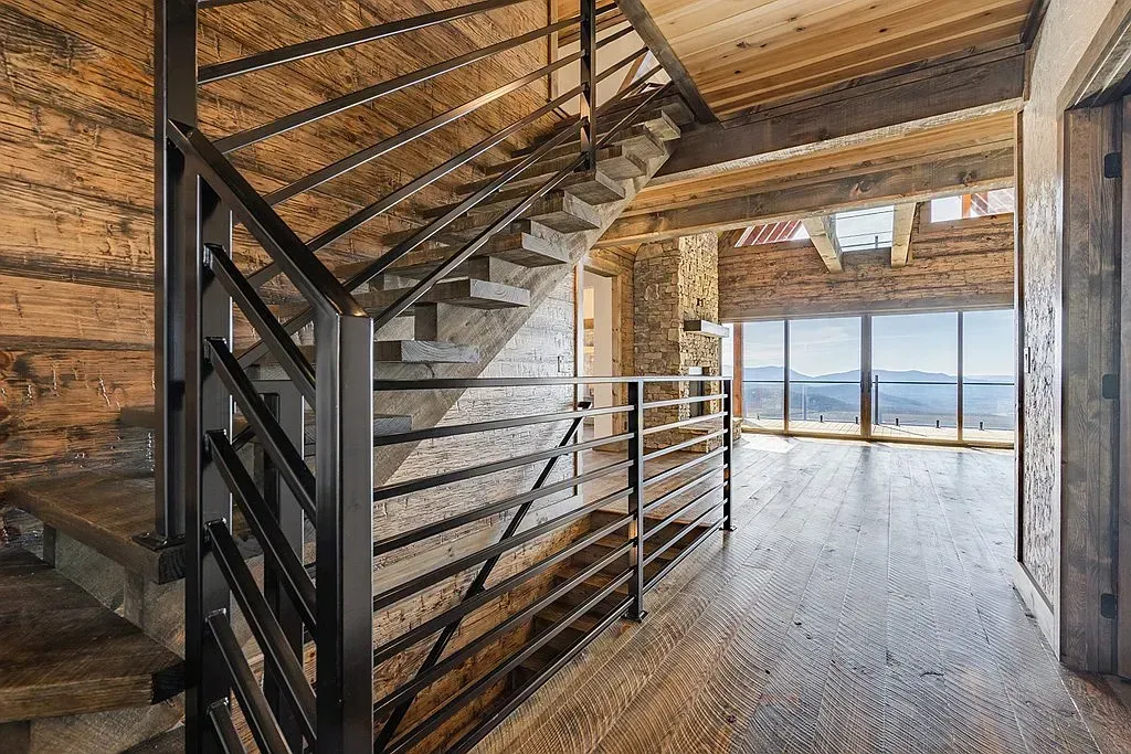 A rustic wooden staircase with a black metal railing leads to a hallway overlooking a scenic mountain view.