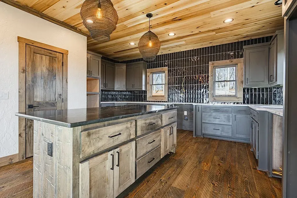 Rustic kitchen with wooden cabinets, a large central island, dark countertops, and pendant lights under a wood ceiling.