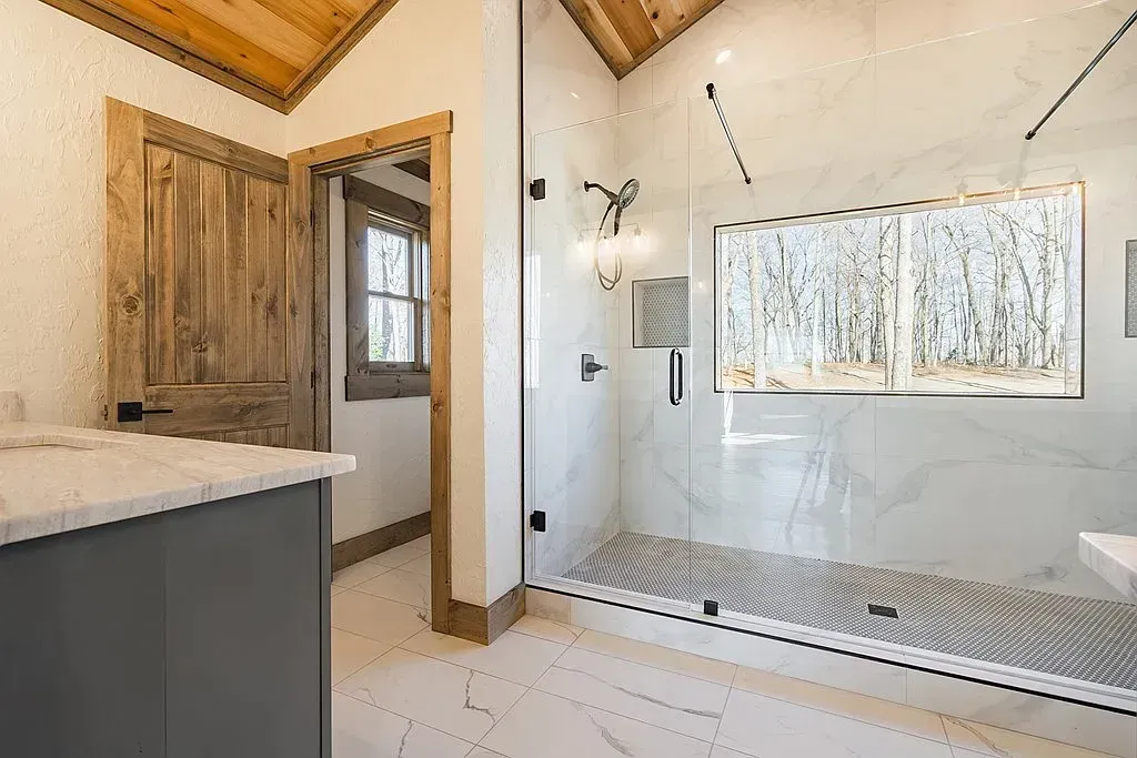 Modern bathroom with a large glass walk-in shower, white marble-style walls, grey vanity, and a wooden ceiling.