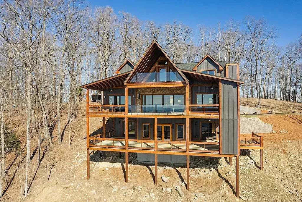 A large multi-story wooden cabin on stilts nestled in a hillside forest with bare trees under a clear blue sky.