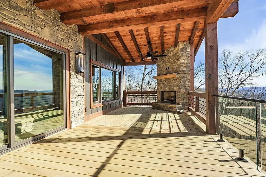 A rustic covered wooden deck with a stone fireplace, large glass doors, and a scenic outdoor view.