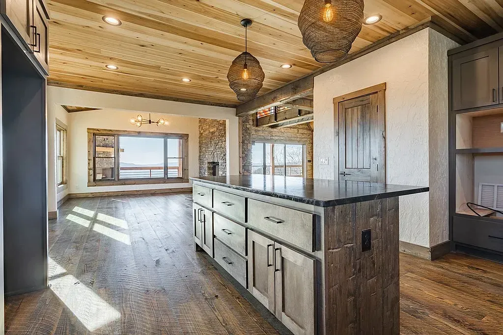 A rustic-style kitchen with a dark wooden island, granite countertop, pendant lights, and a view into a bright dining area.