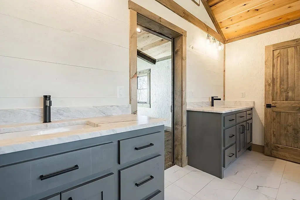 A bathroom with two gray vanities, marble countertops, shiplap walls, a wooden door, and a vaulted wood-paneled ceiling.