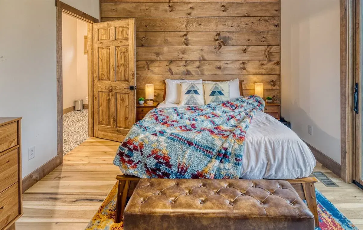 A bedroom with a rustic wood-plank wall, a bed with a colorful patterned quilt, and a brown tufted bench in the foreground.