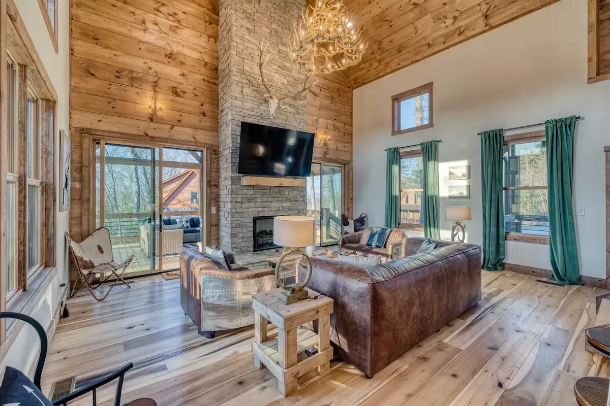A rustic living room with wood paneled walls, a tall stone fireplace, leather furniture, and teal curtains.