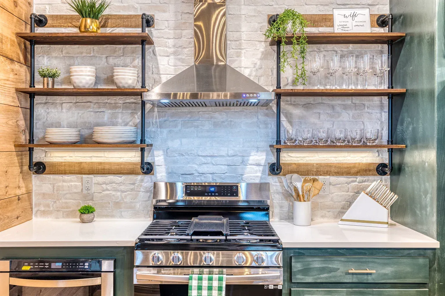 A modern kitchen featuring a stainless steel stove, white brick backsplash, dark green cabinets, and wooden pipe shelving.