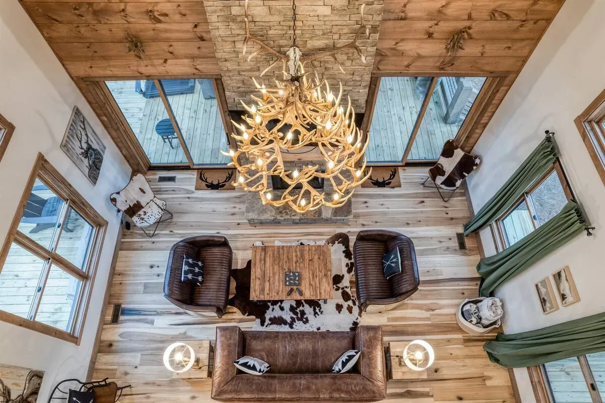 High-angle view of a rustic living room with wood floors, leather furniture, and a large antler chandelier.