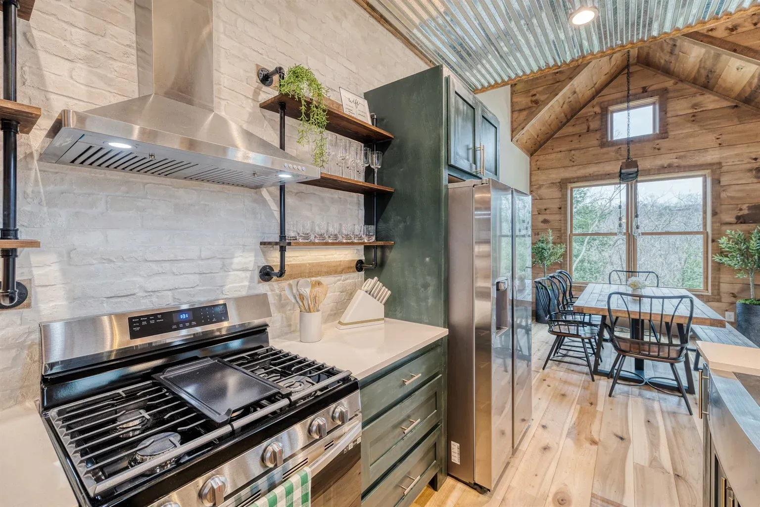 Rustic kitchen with stainless steel stove, white brick wall, green cabinets, and dining area viewed through a doorway.
