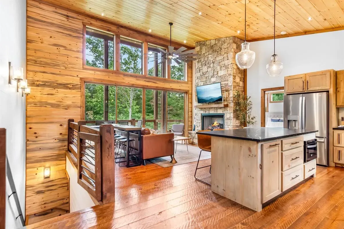 A rustic, open-concept living space with wood walls, vaulted ceilings, a stone fireplace, and a kitchen island.