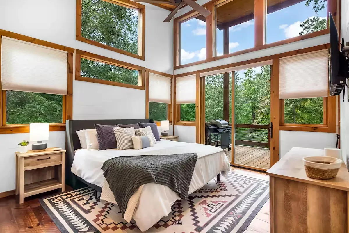 A bedroom with a king bed, patterned rug, wooden nightstand, and large floor-to-ceiling windows looking out onto trees.