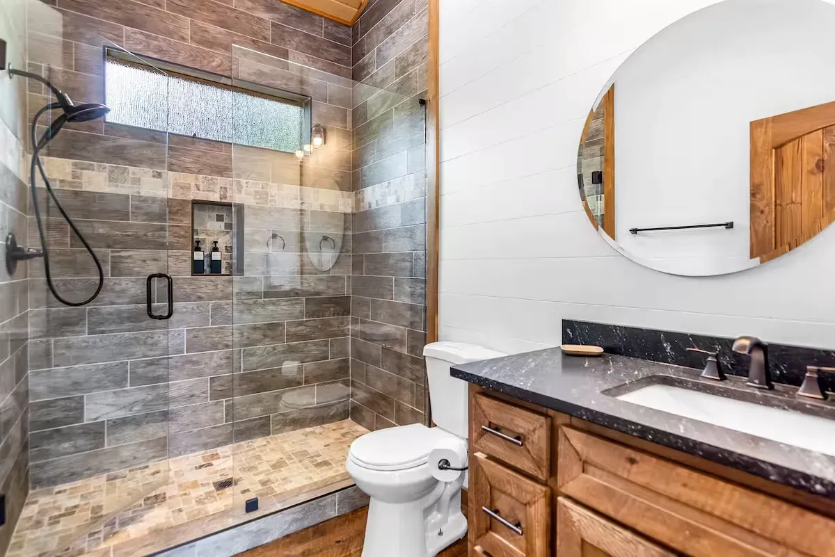 A bathroom with a tiled walk-in shower, a white toilet, and a wood vanity with a dark countertop and round wall mirror.