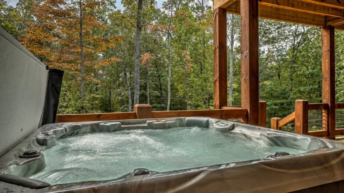 A bubbling hot tub on a wooden deck overlooking a forest with lush green and fall-colored trees.