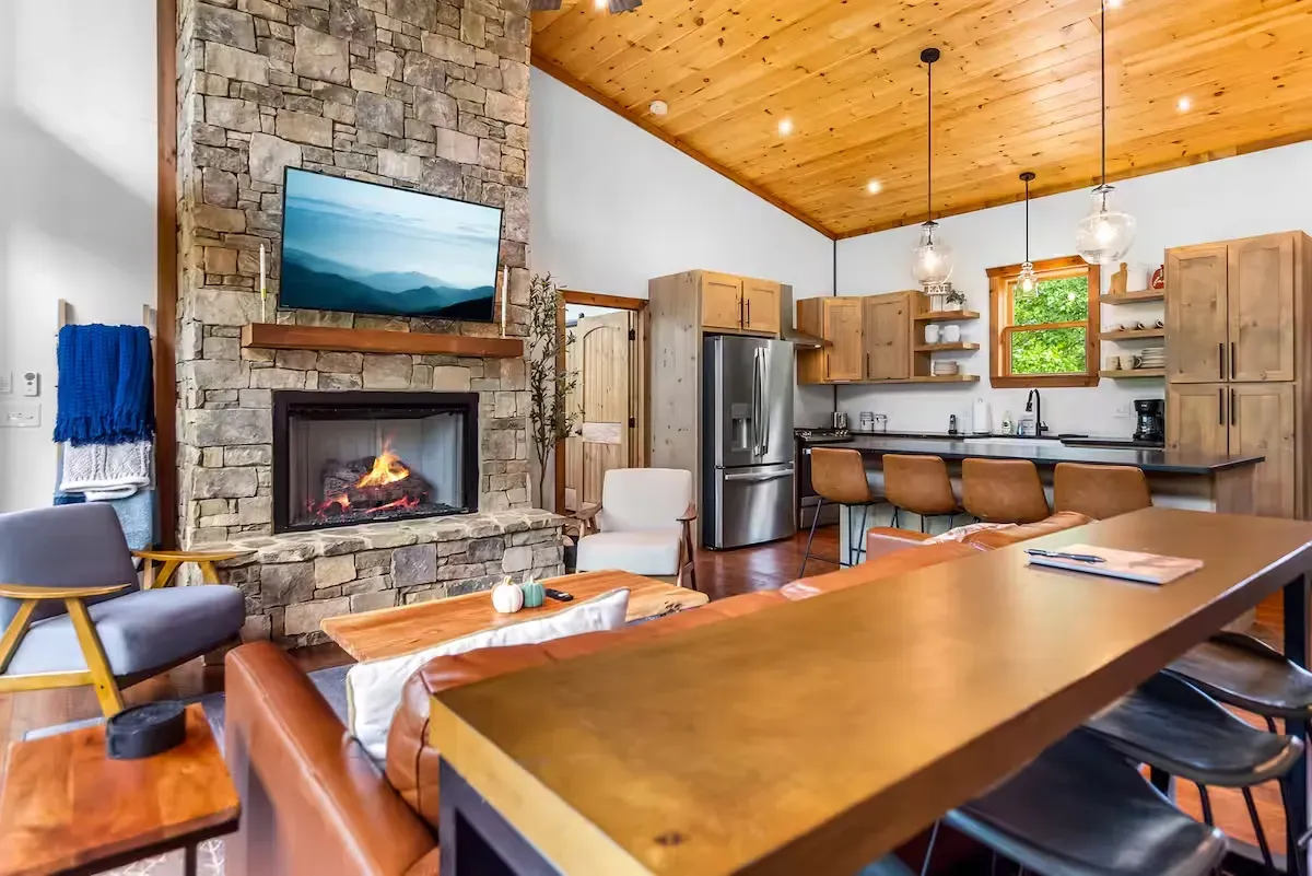 A spacious cabin interior with a stone fireplace, leather sofa, kitchen island with seating, and wood-paneled ceiling.
