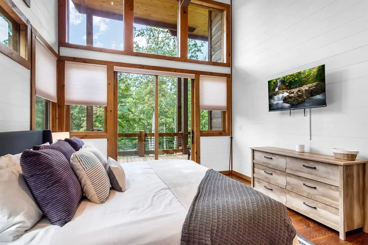 A sunlit bedroom features a bed with purple and patterned pillows facing a wood dresser and a wall-mounted TV.