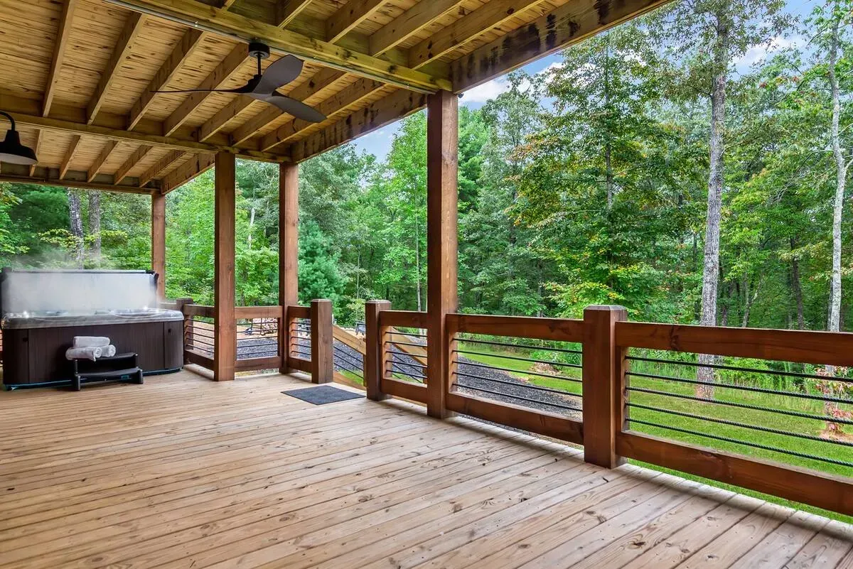 A covered wooden deck featuring a hot tub and cable railings, overlooking a lush green forest.