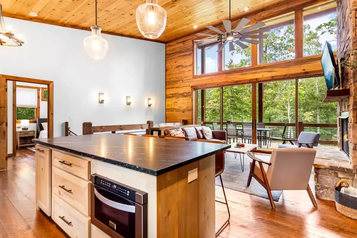 A modern kitchen island with a built-in microwave faces an open-concept living area with large windows overlooking trees.