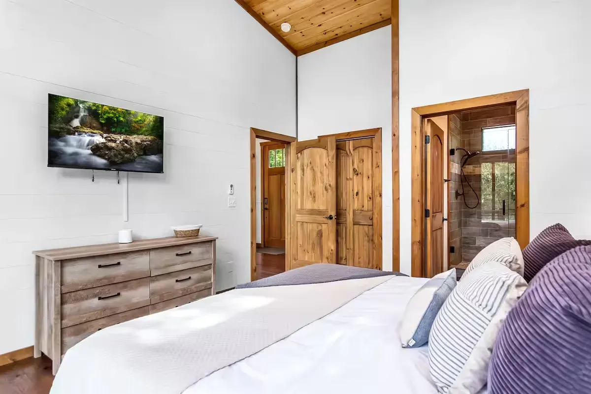 A bedroom with a wooden dresser, wall-mounted television, and a king-sized bed, featuring rustic wood doors and trim.