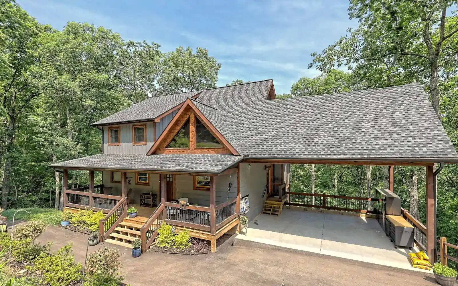 A rustic two-story house with a wooden wraparound porch and an attached carport, nestled in a lush, green forest setting.