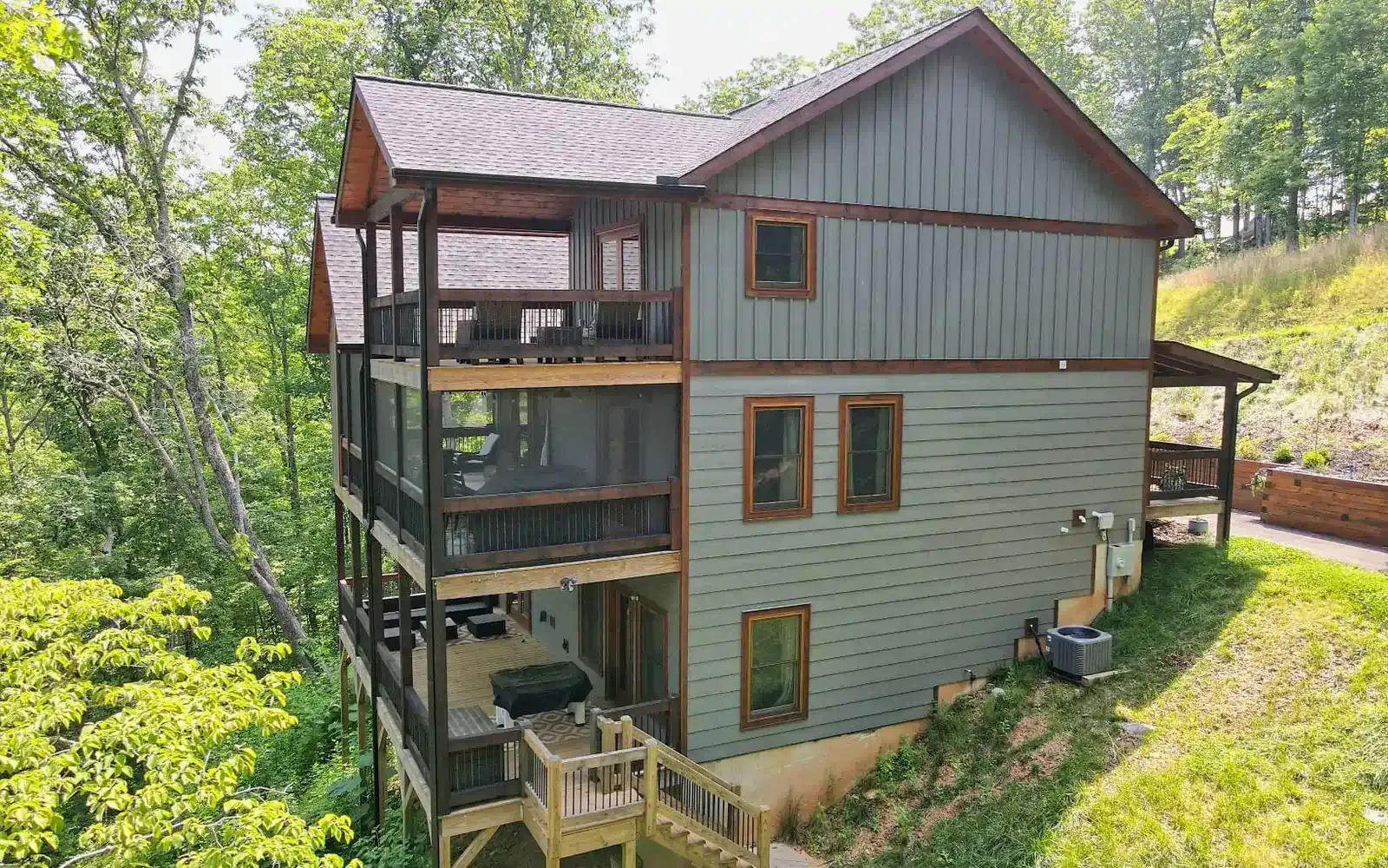 A three-story rustic cabin with gray horizontal siding and a tiered screened-in porch, surrounded by lush green trees.