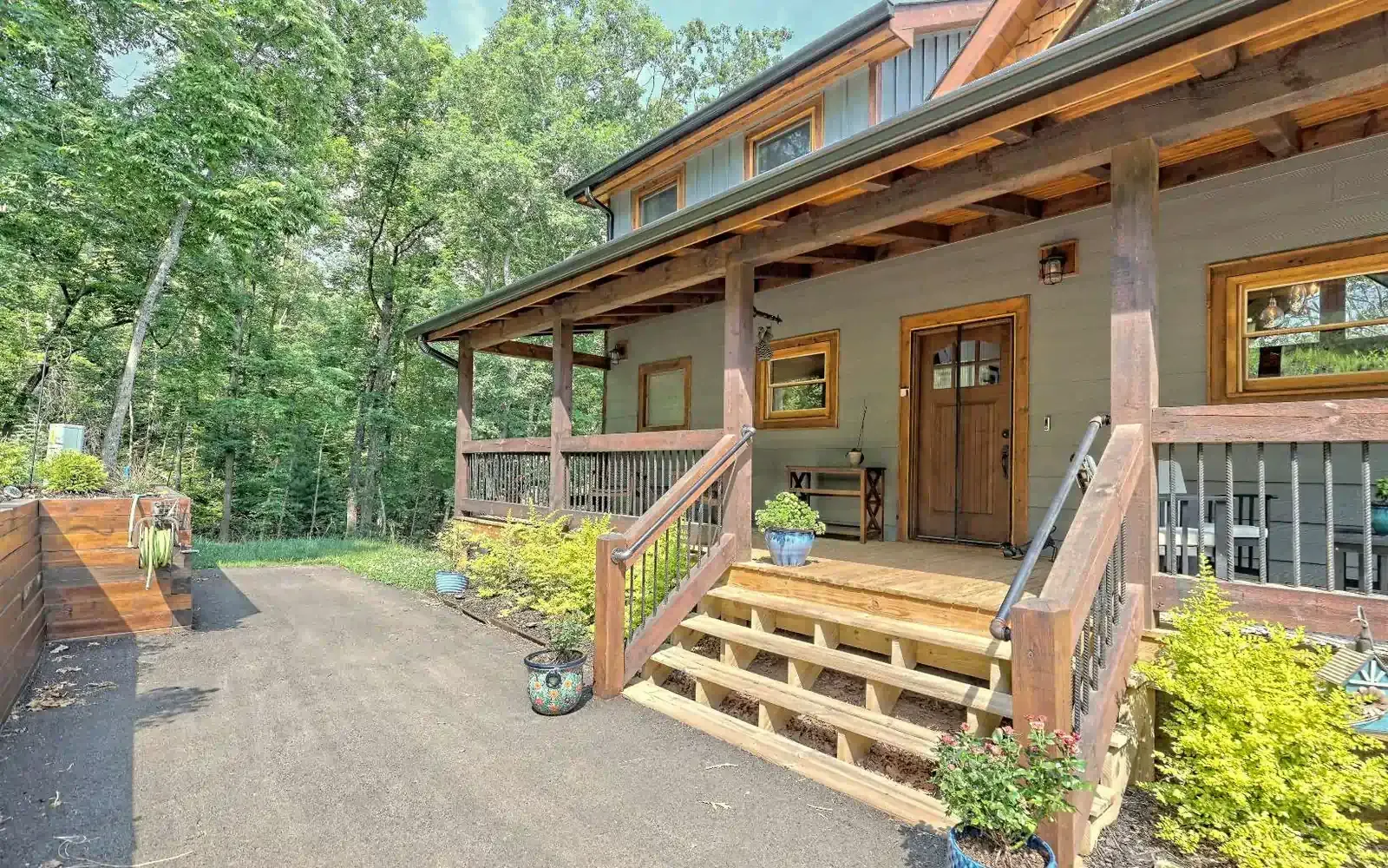 A wooden cabin with a spacious front porch, stairs, and a gravel driveway, surrounded by lush green trees.