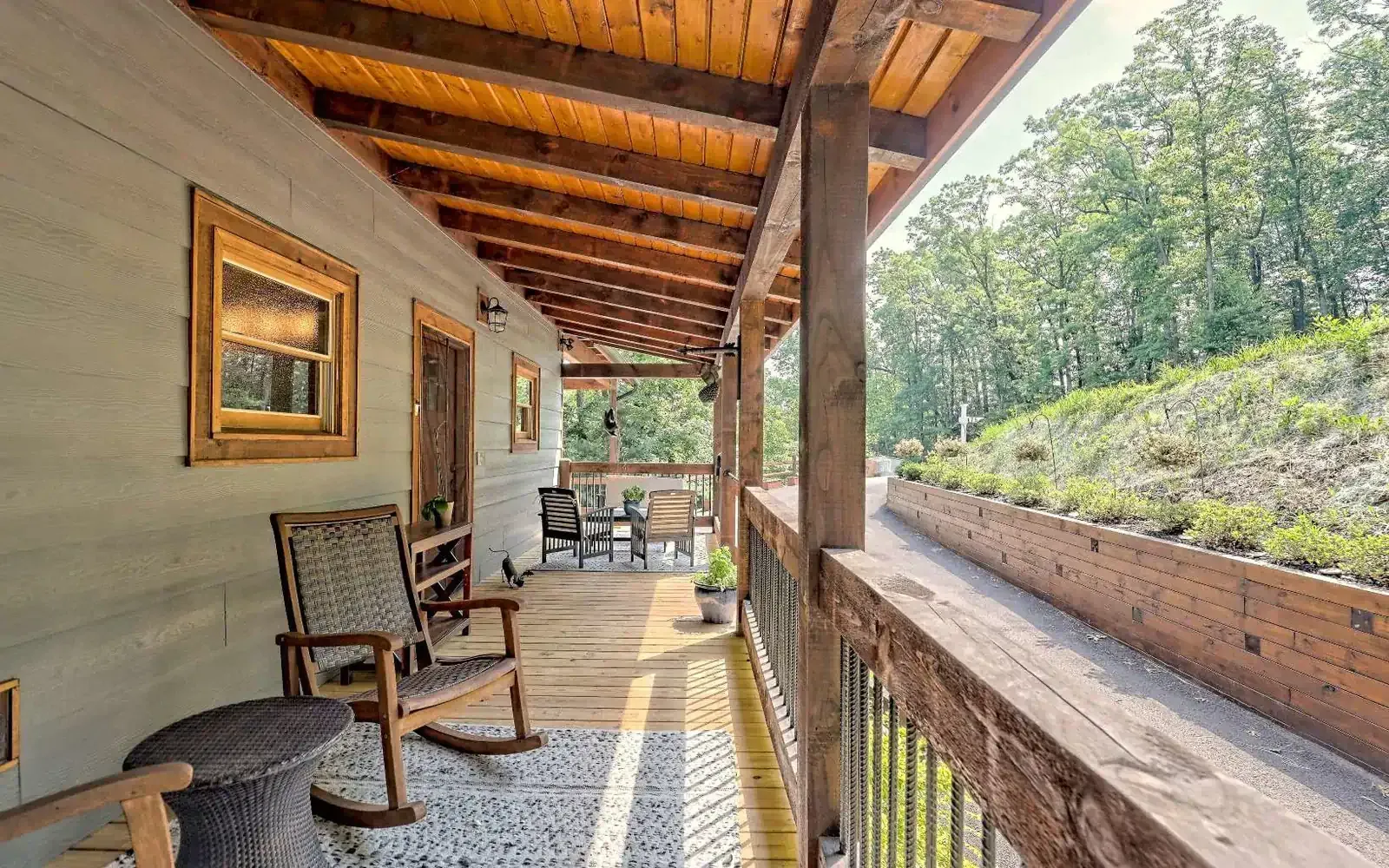 A wooden porch featuring rocking chairs, a rustic ceiling, and railings, overlooking a grassy hill and trees.