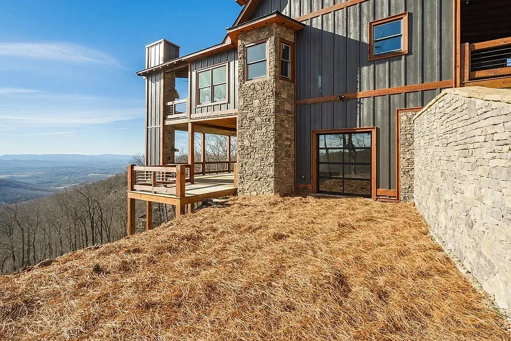 A modern home with wood siding and stone accents sits on a hill overlooking a vast, forested landscape under a blue sky.
