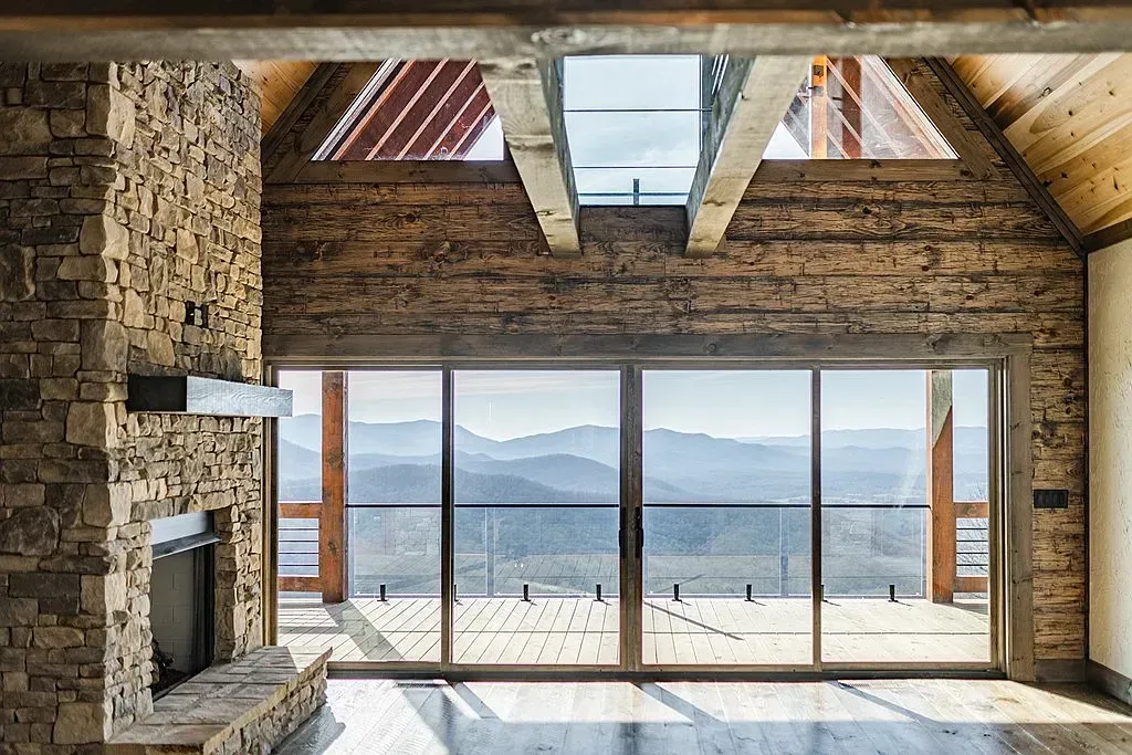 A rustic living room with a stone fireplace, exposed wooden beams, and a large sliding glass door overlooking mountains.