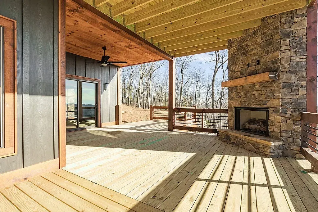 A spacious covered wooden deck features a stone outdoor fireplace and views of a wooded area through a glass door.