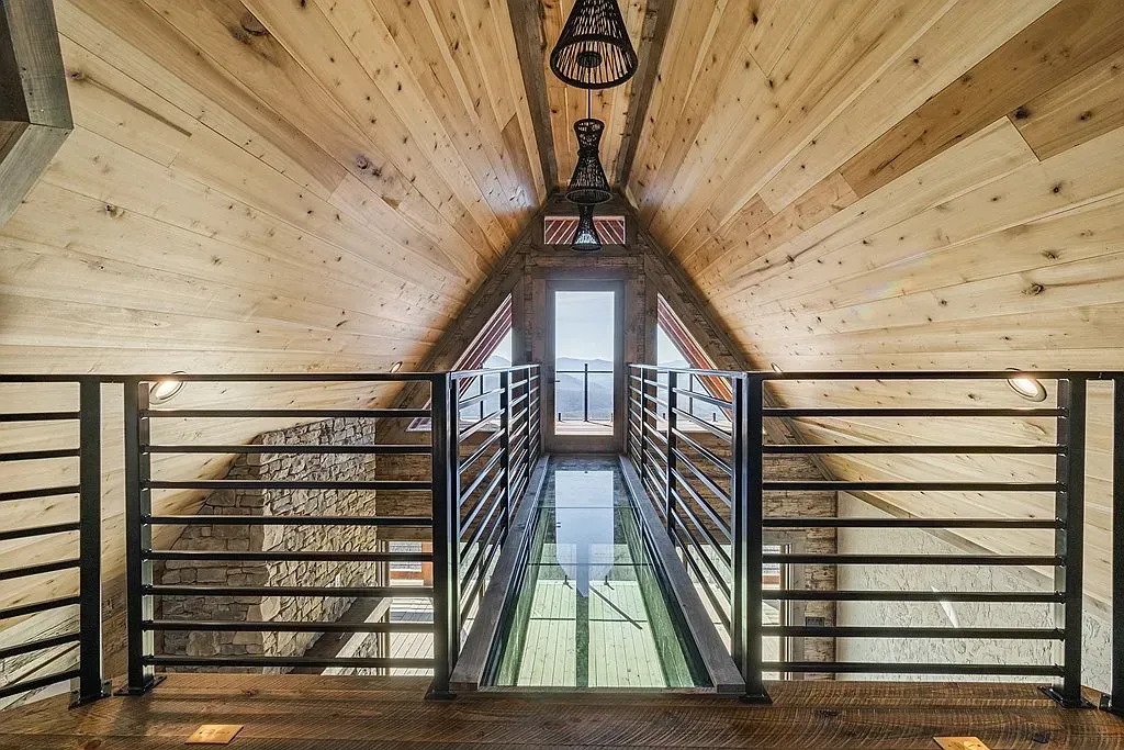 An A-frame wooden loft features a glass-floor walkway leading to a window, flanked by black metal railings and light fixtures.