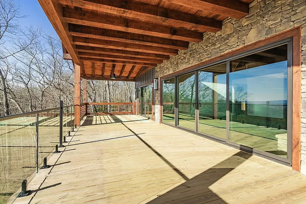 A wooden deck with a glass railing under a timber-framed porch, reflecting trees in large sliding glass doors.