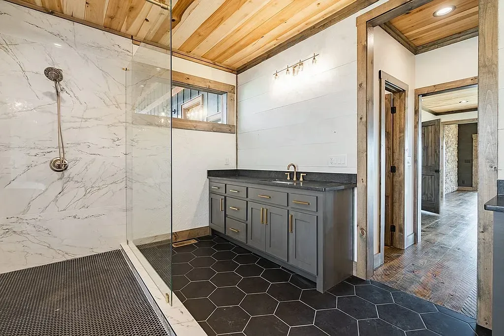 A bathroom with a dark gray vanity, a glass-walled shower with white marble walls, and black hexagonal floor tiles.