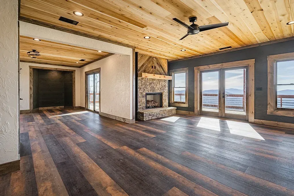 An open, rustic living room featuring wide-plank wood floors, a natural stone fireplace, and light wood paneled ceilings.