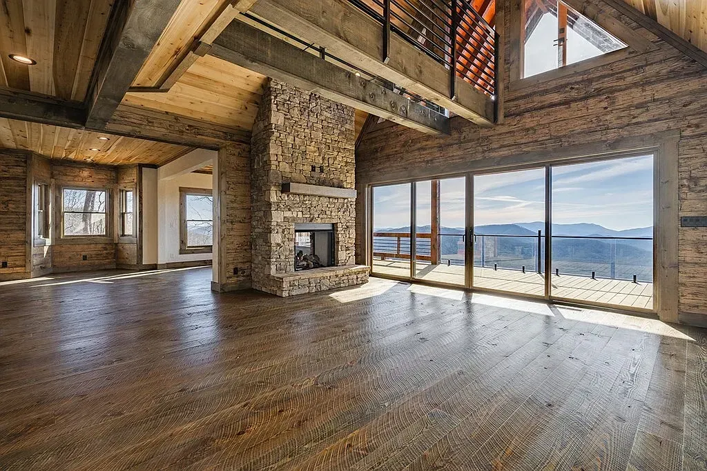 A rustic, high-ceilinged living room with stone walls, hardwood floors, a large central fireplace, and mountain views.