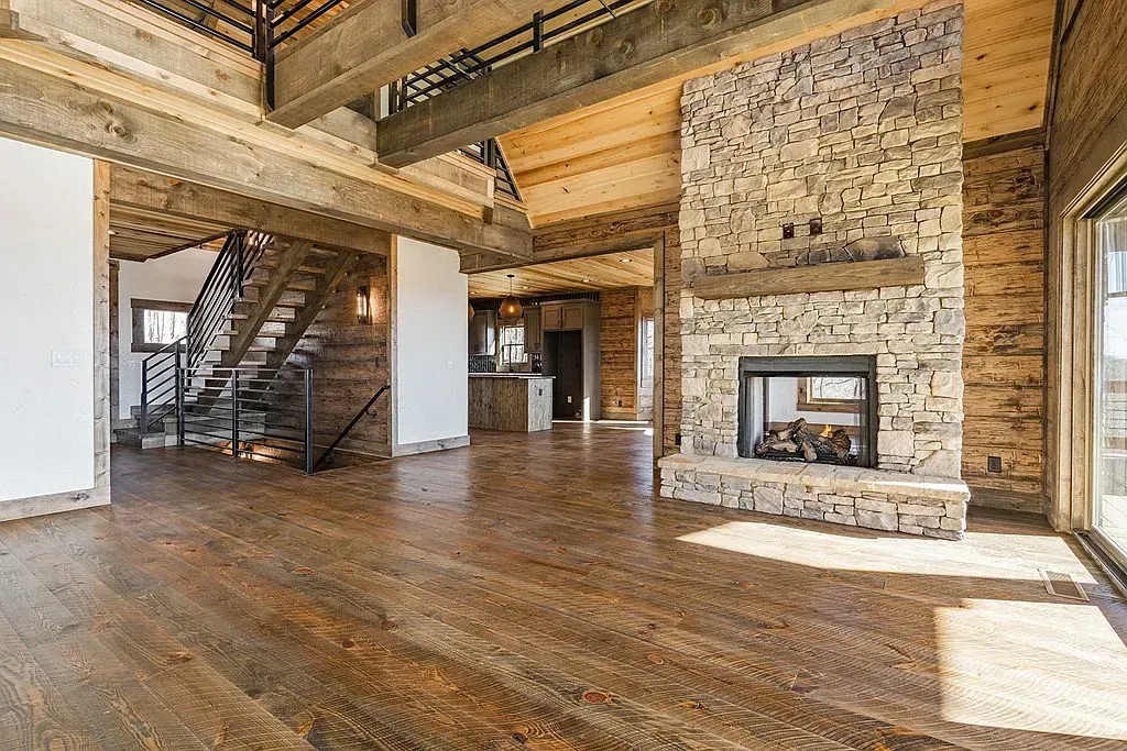 A rustic interior with dark wood floors, exposed ceiling beams, a large stone fireplace, and a staircase in the background.