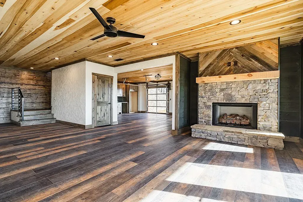 Rustic living room with wood-plank ceiling, dark wood floors, stone fireplace, and open doorway leading to another room.