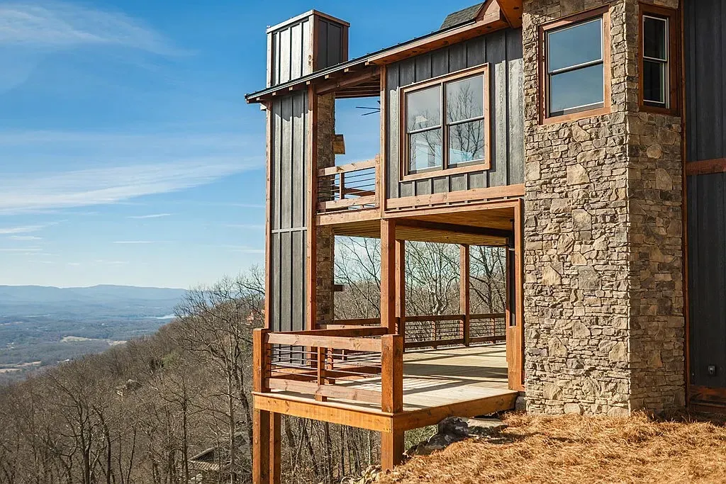 A modern home with wood, stone, and metal siding features a multi-level deck overlooking a vast, hilly landscape.