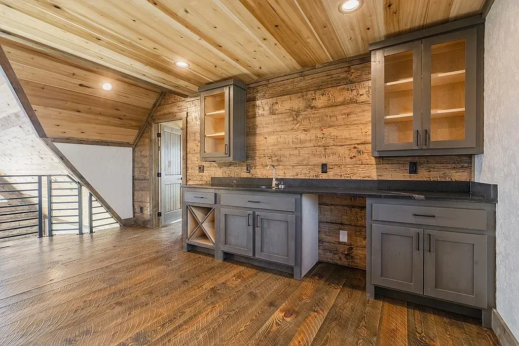 Rustic wet bar with gray cabinets, wood-paneled ceiling, and brick wall in a finished basement with stairs.