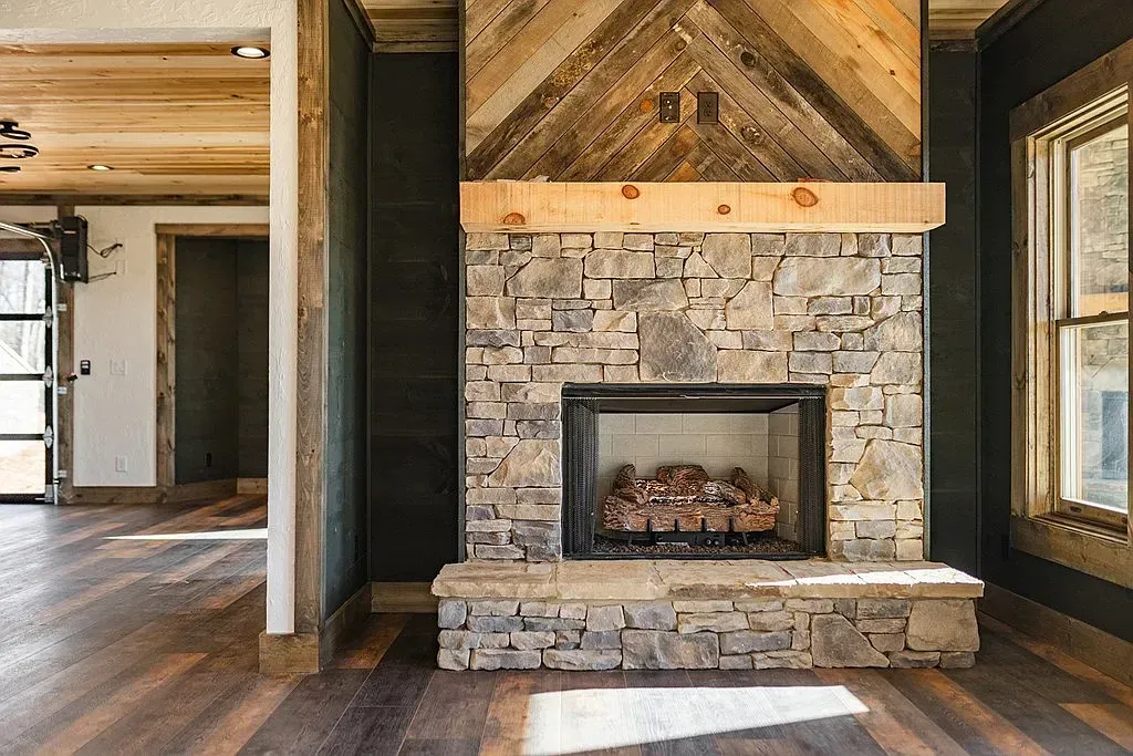 A rustic stone fireplace with a thick wood mantle and chevron-patterned wood paneling above in a modern cabin interior.