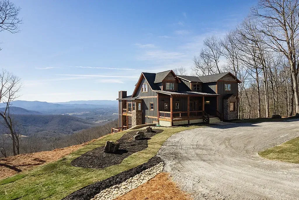 A multi-story cabin with a stone foundation and wood siding sits on a grassy hillside overlooking distant mountains.