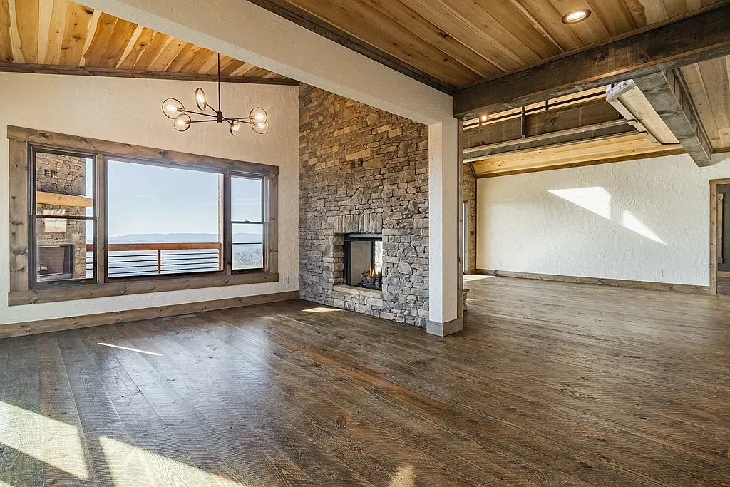 An empty living room features a stone fireplace, large windows, wood-paneled ceilings, and dark wood flooring.