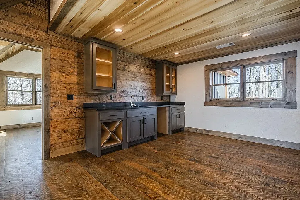 A room with wood-paneled walls and ceiling, dark cabinetry with wine storage, and a window overlooking a wooded area.