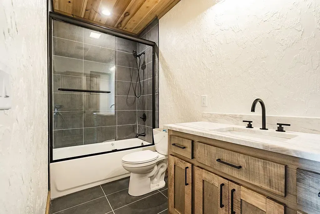 A rustic-style bathroom with a vanity, toilet, and a bathtub with a glass shower door and dark-tiled floor.